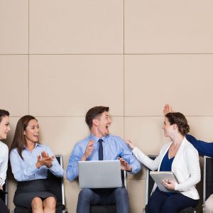 Five cheerful business people sitting in row at wall in waiting room. Young businessman with laptop is cheering ecstatically. Others are congratulating him and smiling.