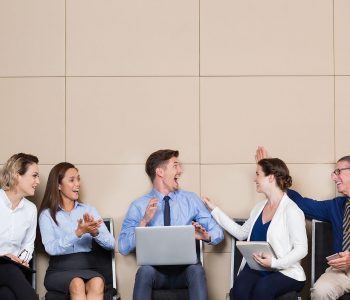 Five cheerful business people sitting in row at wall in waiting room. Young businessman with laptop is cheering ecstatically. Others are congratulating him and smiling.