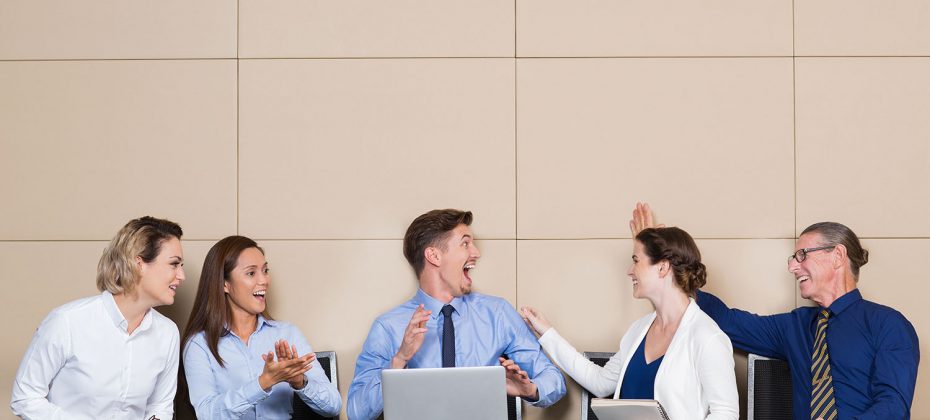 Five cheerful business people sitting in row at wall in waiting room. Young businessman with laptop is cheering ecstatically. Others are congratulating him and smiling.